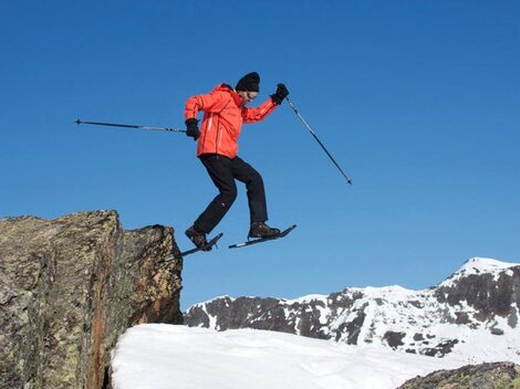 Ein Mann in leuchtend orangefarbener Jacke springt über einen Felsen im Schnee. Im Hintergrund sind schneebedeckte Berge und ein klarer blauer Himmel zu sehen.
