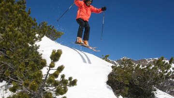 Ein Skifahrer springt freudig über einen Schneehang. Im Hintergrund sind verschneite Berge und blauer Himmel zu sehen.