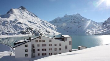 Ein schneebedecktes Bergpanorama mit einem ruhigen See im Vordergrund. Im Vordergrund steht ein modernes Gebäude mit großen Fenstern.