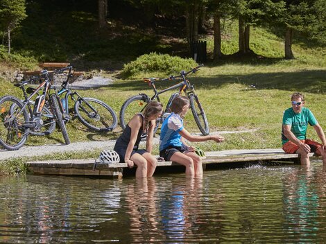 Two girls are sitting by the shore of a calm pond, dipping their feet in the water. In the background, there are bicycles standing among the trees. | © TVB Paznaun - Ischgl