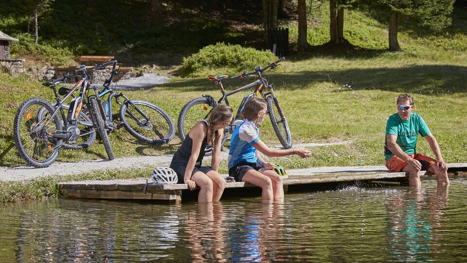 Zwei Mädchen sitzen am Ufer eines ruhigen Teiches und tauchen ihre Füße ins Wasser. Im Hintergrund stehen Fahrräder zwischen den Bäumen. | © TVB Paznaun - Ischgl