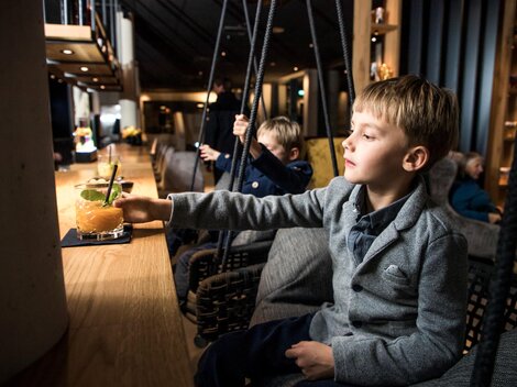 A boy in a plain gray suit is sitting at the bar and reaching for a drink. In the background, other children and a cozy interior are visible.
