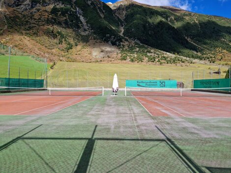 A tennis court in a picturesque mountain landscape. The surroundings are green and sunny with a clear blue sky.