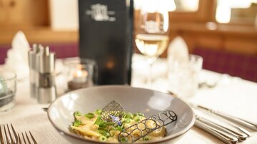 Ein stilvoll gedeckter Tisch mit einem Teller Pasta und frischem Gemüse. Im Hintergrund sind ein Getränk und eine Menükarte zu sehen.
