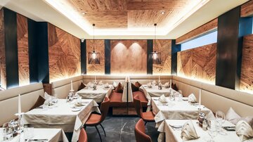 Ein elegantes Restaurant mit schön gedeckten Tischen und modernem Interieur. Die Holzdecke und die sanfte Beleuchtung schaffen eine angenehme Atmosphäre.