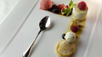 Ein kunstvoll angerichtetes Dessert mit verschiedenen Eiskugeln, frischen Beeren und einer cremigen Zutat. Ein silberner Löffel ergänzt die elegante Präsentation auf dem weißen Teller.