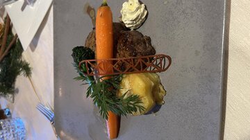 Ein kunstvoll angerichteter Teller mit Fleischbällchen, einer Karotte, Brokkoli und einer Kartoffelbeilage. Oben darauf befindet sich eine kleine Portion Sahne.