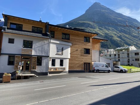A modern building with wood cladding and large windows in a mountain landscape. In the background, a tall mountain is visible under a blue sky.