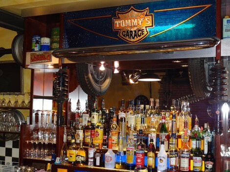 A bar named "Tommy's Garage," filled with various bottles of alcohol and glasses. Tires are hanging on the wall, and the lighting is cozy.