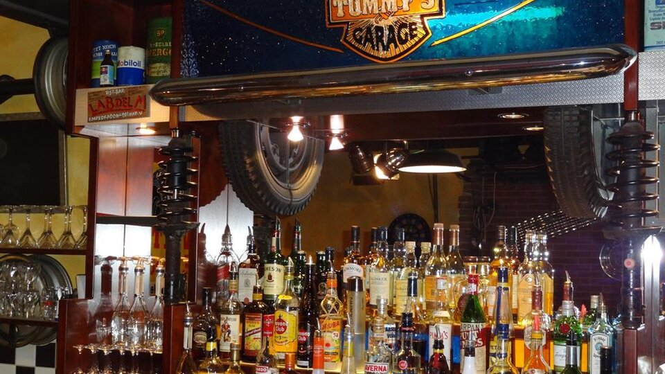 Eine Bar mit dem Namen "Tommy's Garage", voll mit verschiedenen Alkoholflaschen und Gläsern. An der Wand hängen Reifen und die Beleuchtung ist gemütlich.