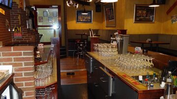 Eine gemütliche Bar mit einer langen Theke und vielen Gläsern. Im Hintergrund sind Tische und Stühle für Gäste zu sehen.