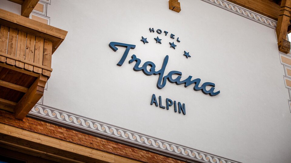 Ein Hotel-Schild mit dem Namen "Trofana Alpin" ist an der Wand angebracht. Die Fassade zeigt rustikale Holzelemente und eine elegante Schriftart.