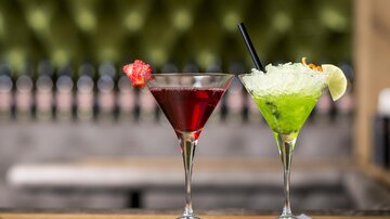 Zwei farbenfrohe Cocktails stehen auf einem Tisch. Links ein roter Cocktail mit einer Fruchtdekoration, rechts ein grüner Cocktail mit einer Limettenscheibe.