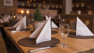 Ein eleganter Tisch in einem Restaurant, schön gedeckt mit Stoffservietten und Gläsern. Im Hintergrund sind warme Lichtakzente und eine gemütliche Atmosphäre zu sehen.