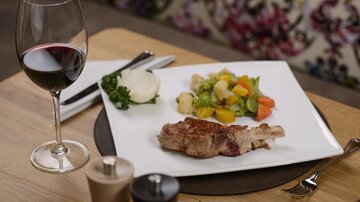 Ein eleganter Teller mit einem Stück Fleisch, buntem Gemüse und einem Klecks Kartoffelpüree. Dazu ein Glas Rotwein auf einem Holztisch.