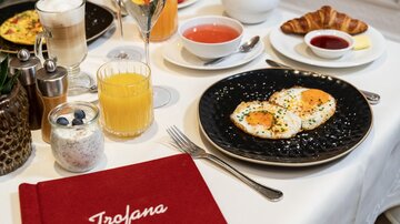 Ein schön gedeckter Tisch mit einem Frühstücksarrangement. Dazu gehören Spiegeleier, Croissants, Säfte und Joghurt.