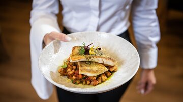 Ein gut angerichteter Teller mit gebratenem Fisch und buntem Gemüse. Der Service wird von einer Person in weißem Hemd präsentiert.