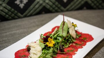 Ein elegantes Gericht mit rotem Carpaccio, umgeben von frischem Salat und essbaren Blüten. Die Präsentation ist ansprechend auf einem weißen Teller arrangiert.