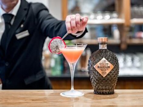Ein Barkeeper stellt einen bunten Cocktail mit einer Fruchtdekoration auf die Theke. Neben dem Getränk steht eine Flasche mit dem Etikett "BAIGUR".