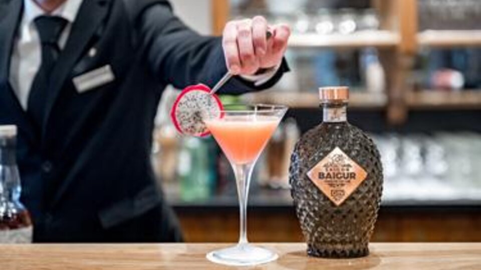 Ein Barkeeper stellt einen bunten Cocktail mit einer Fruchtdekoration auf die Theke. Neben dem Getränk steht eine Flasche mit dem Etikett "BAIGUR".