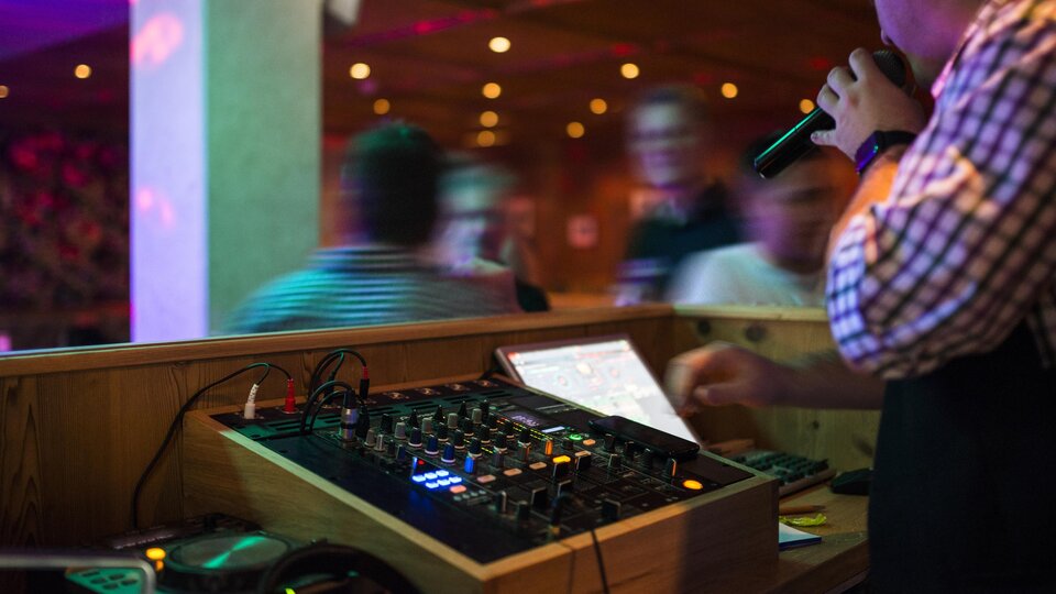 Ein DJ steht hinter einem Mischpult und hält ein Mikrofon in der Hand. Die bunten Lichter und die tanzende Menge schaffen eine lebendige Partystimmung.