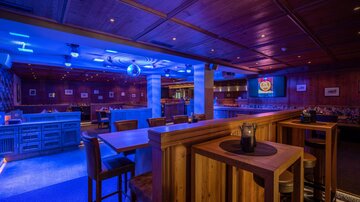 Eine stilvolle Bar mit Holzinterieur und blauer Beleuchtung. Es gibt hohe Tische und eine entspannte Atmosphäre.