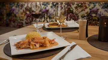Ein elegantes Essen mit Garnelen und gelbem Reis auf einem weißen Teller. Im Hintergrund sind Weingläser und eine dekorative Wand zu sehen.