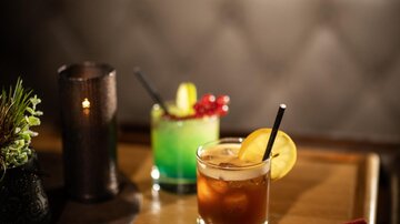 Zwei bunte Cocktails auf einem Tisch, einer mit einer orangefarbenen Garnitur und der andere mit einer grünen Farbe. Daneben liegt eine rote Getränkekarte und eine Kerze sorgt für eine gemütliche Atmosphäre.
