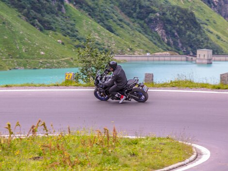 A motorcyclist rides along a winding road next to a clear lake. The green landscape around him creates an impressive backdrop.