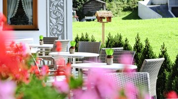 Ein einladender Außenbereich mit Tischen und Stühlen. Bunte Blumen umrahmen die Szene und der grüne Rasen sorgt für eine fröhliche Atmosphäre.