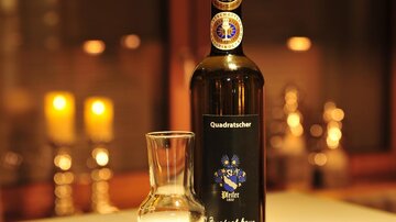 Eine Flasche Obstler steht auf einem Tisch. Daneben sind mehrere Gläser und im Hintergrund leuchten Kerzen.