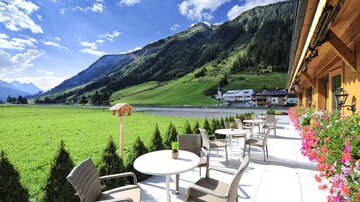 Eine wunderschöne Terrasse mit Blick auf grüne Wiesen und Berge. Im Hintergrund sind kleine Häuser und ein blauer Himmel zu sehen.