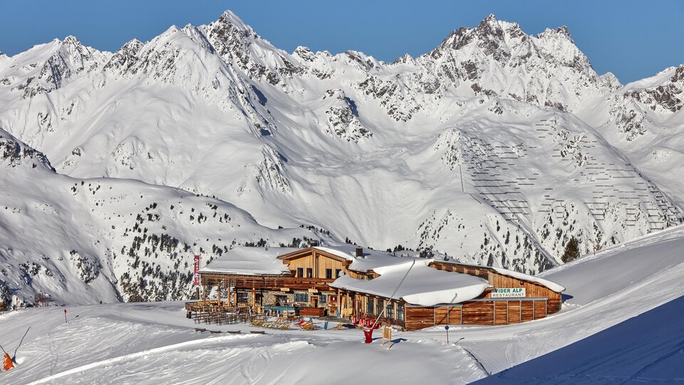 Eine gemütliche Berghütte liegt in einer schneebedeckten Landschaft. Im Hintergrund ragen majestätische Berge unter einem klaren Himmel empor.