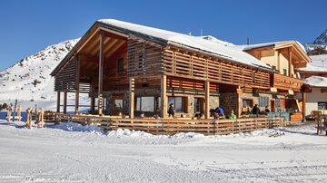Ein gemütliches Holzgebäude im Schnee, umgeben von Bergen. Es gibt Menschen, die draußen sitzen und die Winterlandschaft genießen.