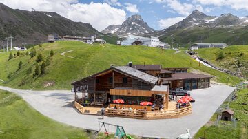 Eine gemütliche Berghütte umgeben von grünen Hügeln und beeindruckenden Bergen. Die Hütte hat eine Terrasse mit rot-weißen Sonnenschirmen.