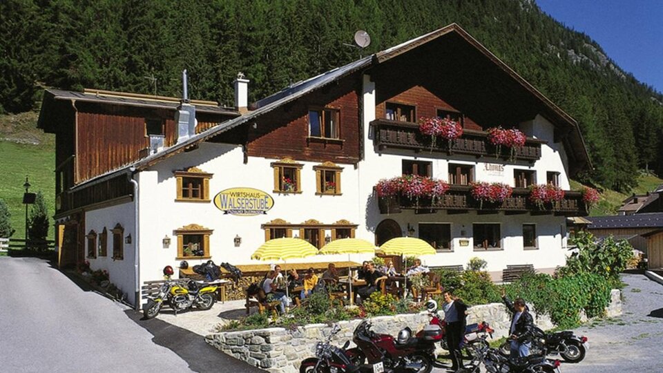 Ein gemütliches Gasthaus in den Bergen mit blühenden Balkonen und Sonnenschirmen. Vor dem Gebäude parken mehrere Motorräder.