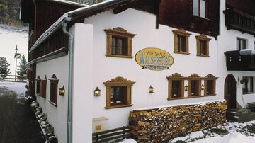 Ein traditionelles Gasthaus mit weißer Fassade und schönen Holzdetails. Im Vordergrund sieht man einen Holzstapel und winterliche Landschaft im Hintergrund.