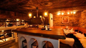 Eine gemütliche Weinstube mit einem Holztheke und sanfter Beleuchtung. Die Wände sind aus Stein und für eine warme Atmosphäre sorgen weiche Felle auf den Sitzen.