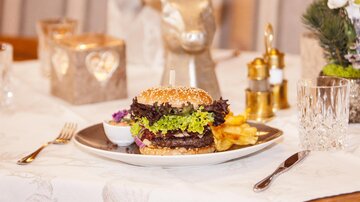Ein köstlicher Burger auf einem Teller mit frischem Salat und Fritten. Der Tisch ist elegant gedeckt, mit Kerzen und dekorativen Elementen im Hintergrund.
