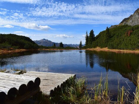 A tranquil lake surrounded by mountains and forest. The sky is clear with a few clouds.