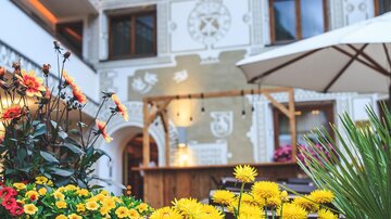 Ein schöner Innenhof mit blühenden Blumen und einem Gasthof im Hintergrund. Die Wand ist kunstvoll gestaltet und lädt zum Verweilen ein.