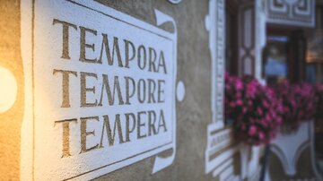 Eine Wand mit der Aufschrift "TEMPORA TEMPORE TEMPERA". Im Hintergrund sind pinke Blumen zu sehen.