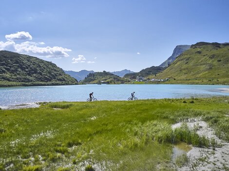Two cyclists ride along a sparkling lake. In the background, green hills and a blue sky are visible.