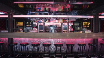 Eine stilvolle Bar mit einer langen Holztheke und modernen Sitzgelegenheiten. Die Beleuchtung ist in sanften Pinktönen gehalten und schafft eine einladende Atmosphäre.