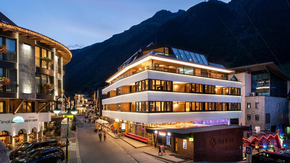 Ein modernes Gebäude in einer malerischen Alpenstadt bei Dämmerung. Im Hintergrund sind hohe Berge und eine belebte Straße mit Lichtern zu sehen.