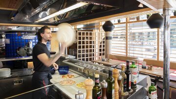 Ein Koch in einer modernen Pizzeria wirft einen Pizzateig in die Luft. Auf der Arbeitsfläche sind verschiedene Gewürze und Zutaten sichtbar.
